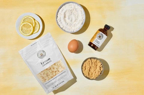Assorted ingredients for Chewy Lemon Blondies on a yellow surface, including lemon extract, slices of lemon, lemon crumbles, an egg, and two types of sugar in small bowls